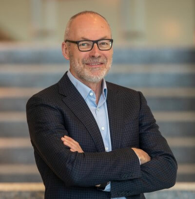 Dr. Pinniger in glasses, arms crossed and a blue collared shirt with black suit jacket.